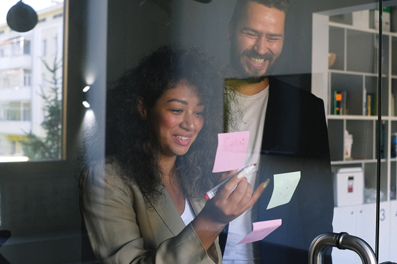 Two cowrkers smiling whilst brainstorming using post-it notes on a glass wall